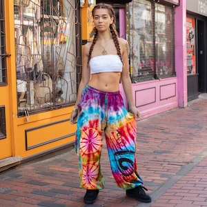 Op de afbeelding: Een vrouw draagt een wit crop top en een kleurrijke tie-dye broek met een bloemenpatroon. De broek heeft wijde pijpen en is gemaakt van een zachte, vloeiende stof.