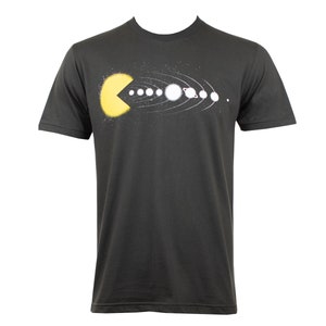 Puede incluir: Una camiseta gris oscuro con un Pac-Man amarillo comiendo los planetas del sistema solar, incluido el sol, en una impresión gráfica blanca.