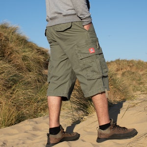 May include: A pair of green cargo shorts with a red logo on the right leg. The shorts are worn by a person standing on a sandy beach.