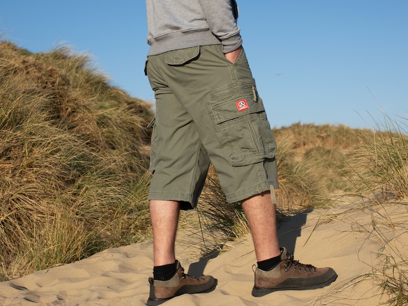 May include: A pair of green cargo shorts with a red logo on the right leg. The shorts are worn by a person standing on a sandy beach.