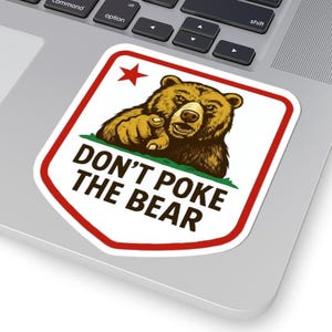 May include: A sticker featuring a brown bear pointing its finger, with the text "DON'T POKE THE BEAR" in bold letters. The sticker has a red border and a red star in the upper left corner. The sticker is placed on a laptop.