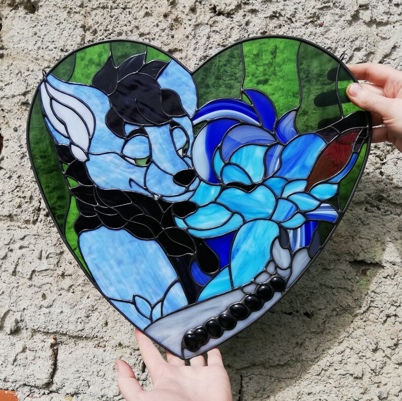 May include: A heart-shaped stained glass art piece featuring two stylized blue and black furry characters. The artwork has a green background and is held by hands against a textured wall.