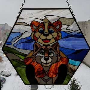 May include: A stained glass panel in a hexagonal shape, featuring two cartoon raccoons. The top raccoon is orange and cream, the bottom one is brown and tan. The background depicts a blue sky and green hills.
