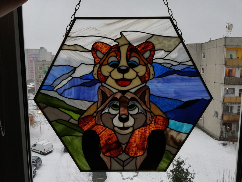 May include: A stained glass panel in a hexagonal shape, featuring two cartoon raccoons. The top raccoon is orange and cream, the bottom one is brown and tan. The background depicts a blue sky and green hills.