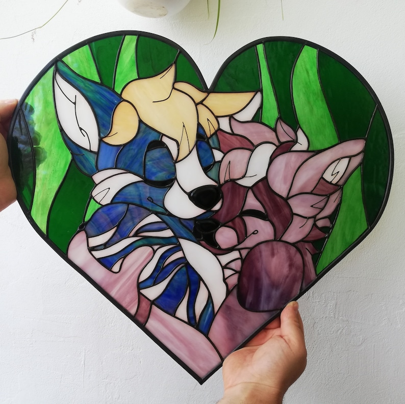 May include: A heart-shaped stained glass art piece featuring two stylized animal figures in shades of blue, pink, and yellow. The artwork has a black border and a green background with vertical lines. The piece is held by hands.