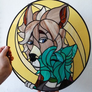 May include: A round stained glass art piece featuring a stylized anthropomorphic wolf with tan and brown fur, blue eyes, and a teal-colored wolf. The artwork is set against a yellow background with black outlines.