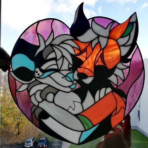 May include: A heart-shaped stained glass art piece featuring two stylized anthropomorphic animals embracing. The artwork uses shades of white, gray, black, orange, and teal, with a pink and purple background. The piece is held up against a window.
