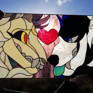 May include: Stained glass panel featuring two stylized animal faces, one cream and the other black and white, with a red heart between them. The panel is held up against the sun, highlighting the colors and details of the artwork.