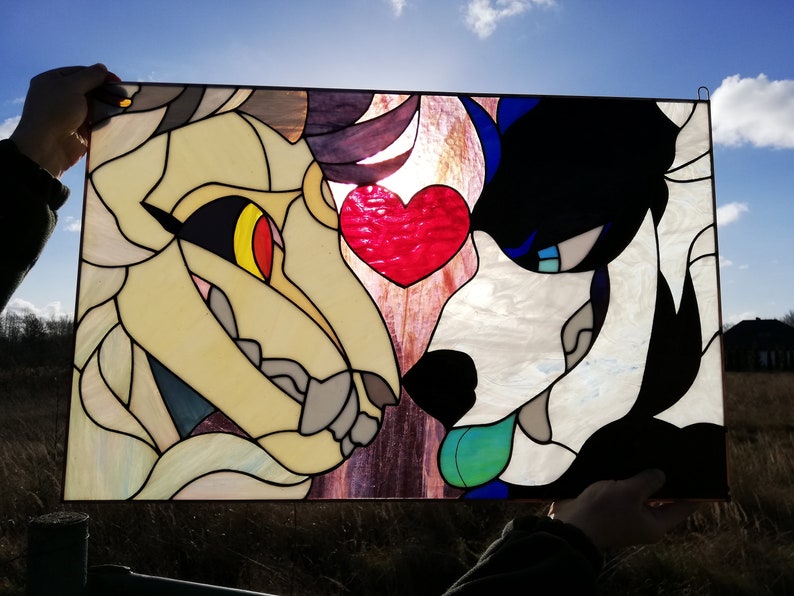May include: Stained glass panel featuring two stylized animal faces, one cream and the other black and white, with a red heart between them. The panel is held up against the sun, highlighting the colors and details of the artwork.