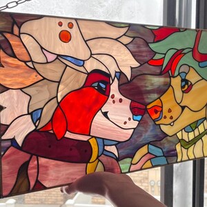 May include: Stained glass panel featuring two anthropomorphic characters with colorful fur and facial features. The artwork is framed with a dark metal border and suspended by a chain. The characters are in shades of red, white, yellow, and green.