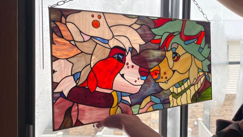 May include: Stained glass panel featuring two anthropomorphic characters with colorful fur and facial features. The artwork is framed with a dark metal border and suspended by a chain. The characters are in shades of red, white, yellow, and green.