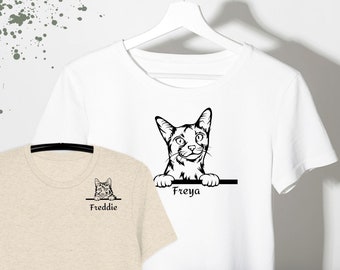 May include: A white t-shirt and a beige t-shirt, both featuring a black line art illustration of a cat peeking over a horizontal line. The white shirt displays the name Freya, while the beige shirt displays the name Freddie.
