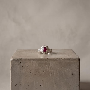 May include: A silver ring featuring a central oval-shaped deep red gemstone. The band of the ring has a textured, organic surface with a series of small, rounded protrusions surrounding the stone, creating a distinct, uneven metal finish.