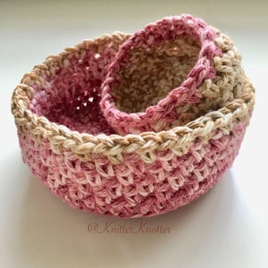 May include: Two crocheted baskets, one nested inside the other. The larger basket is pink and white, while the smaller basket is beige and white. Both baskets have a braided edge.