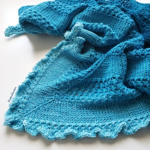 Könnte beinhalten: Ein blauer und türkisfarbener gestrickter Schal mit einem Spitzenmuster. Der Schal ist gefaltet und über eine weiße Oberfläche gelegt.