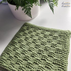 Könnte beinhalten: Ein quadratisches, salbeigrünes Strickstück mit einem strukturierten Korbgeflechtmuster. Der handgefertigte Artikel wird auf einer weißen Oberfläche mit einer Topfpflanze im Hintergrund präsentiert. Der Artikel ist wahrscheinlich ein Waschlappen oder eine kleine Decke.