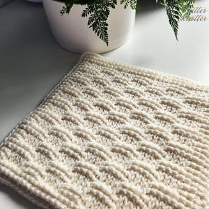 Puede incluir: Cuadrado tejido a mano de color crema con un patrón tejido texturizado. La pieza tejida tiene un diseño de tejido de cesta. Probablemente sea un paño, un paño de cocina o una manta pequeña.