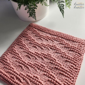May include: A square, knitted item in a dusty rose color. The texture features a repeating pattern of vertical and diagonal lines, creating a textured surface. The item is likely a washcloth or small blanket, with a handmade aesthetic.