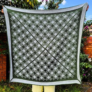 Puede incluir: Una manta de crochet verde y blanca con un patrón de cuadros. La manta está hecha con un hilo suave y tiene un borde con flecos.