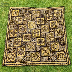 May include: A handmade, square blanket featuring a repeating pattern of geometric shapes in black and gold. The blanket is made of a soft material and is laid out on a green grassy surface.