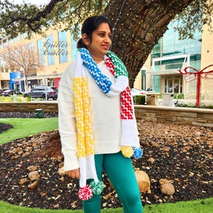 May include: A white crocheted scarf with colorful stripes of red, yellow, blue, green, and white. The scarf has pom-poms on the ends. The scarf is worn by a person standing in front of a tree.