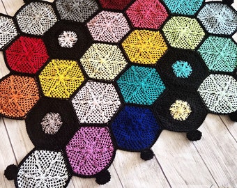 Może przedstawiać: Kolorowy koc szydełkowy z wzorem plastra miodu i czarnymi pomponami na brzegach. Koc wykonany jest z przędzy w różnych kolorach, w tym czerwonym, pomarańczowym, żółtym, zielonym, niebieskim, fioletowym i białym.