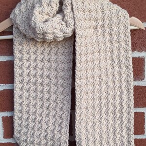 May include: A beige crocheted scarf with a textured pattern. The scarf is folded in half and hanging on a wooden hanger.