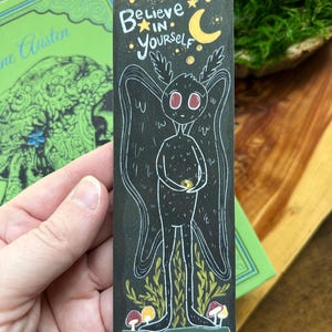 May include: A rectangular black bookmark featuring a white line art illustration of a winged creature with large eyes. The text "Believe in yourself" is written in white above the creature, accompanied by a yellow crescent moon and stars.
