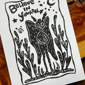 May include: A white rectangular card featuring a black ink block print illustration of a winged creature with horns standing among tall plants. The text "Believe in Yourself" is printed at the top alongside a crescent moon and stars.