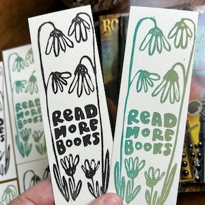 May include: Two rectangular paper bookmarks featuring a hand-drawn floral design with drooping flowers at the top and bottom. One bookmark has black ink, and the other has green ink. Both feature the text "READ MORE BOOKS" in a bold, blocky font.