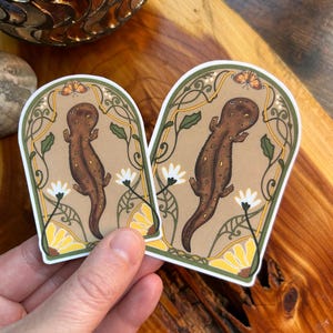 May include: Two die-cut stickers featuring a brown salamander centered within an arched frame. The design includes green vines, white flowers, yellow petals, and a small orange butterfly at the top, all set against a tan background with a white border.