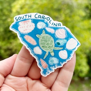 May include: A die-cut sticker shaped like the state of South Carolina with a blue background. The design features a central green sea turtle surrounded by various illustrations of seashells in shades of pink, white, and blue. The text South Carolina is printed in blue along the top edge of the sticker.