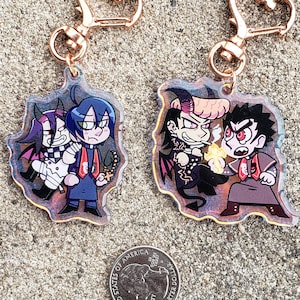 May include: Two colorful character keychains with gold heart-shaped clasps. The keychain on the left features three characters, one with blue hair and a purple and white outfit, one with purple hair and a black and white outfit, and one with black hair and a red and white outfit. The keychain on the right features two characters, one with orange hair and a black and red outfit, and one with black hair and a red and white outfit.