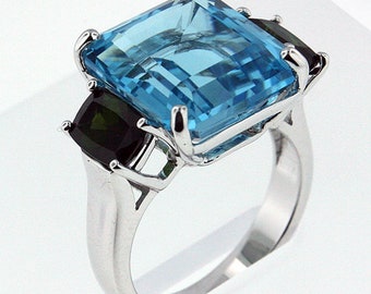 May include: A silver ring with a large, light blue, emerald-cut gemstone and two smaller, dark green, square-cut gemstones.