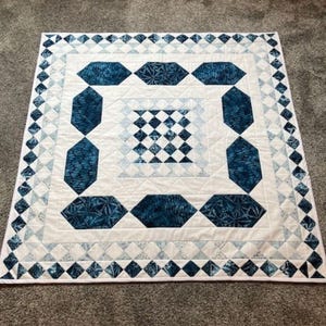 May include: A handmade quilt with a geometric design. The quilt features a white background with a central square of small blue and white squares. Large, dark blue diamond shapes are arranged around the centre, and a border of small triangles completes the design.