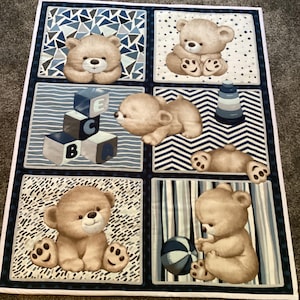 May include: A blue and white fabric panel with six squares featuring brown teddy bears with different expressions and playing with toys. The toys include blocks that spell "ABC", a stacking ring toy, and a beach ball.