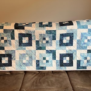 May include: A rectangular quilt featuring a geometric pattern of square blocks in various shades of blue, teal, and white. The design consists of nested squares and solid colour patches arranged in a grid, finished with a white border and stitching.