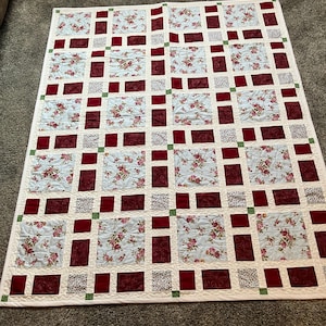 May include: A rectangular quilt featuring a grid pattern of alternating squares. The design includes light blue fabric with a pink floral print, solid deep red squares, and smaller white squares with a subtle grey pattern, accented by tiny green corner squares.