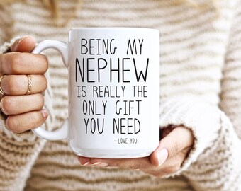May include: White ceramic mug with the text "BEING MY NEPHEW IS REALLY THE ONLY GIFT YOU NEED - LOVE YOU" in black. The mug is held by a person wearing a cream-colored sweater.