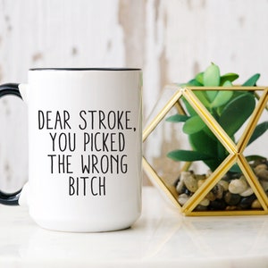 May include: White ceramic mug with a black handle and rim. The mug features the text "DEAR STROKE, YOU PICKED THE WRONG BITCH" in black lettering. A geometric gold terrarium with a faux succulent plant sits beside the mug.