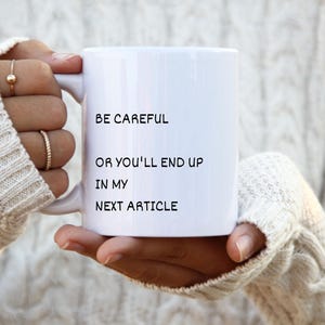 May include: A white ceramic mug featuring the black printed text BE CAREFUL OR YOU'LL END UP IN MY NEXT ARTICLE. The mug has a standard handle and a smooth cylindrical body, designed for holding hot or cold beverages in a simple, functional style.