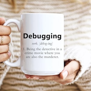 May include: White ceramic mug with the word "Debugging" in a bold black font. The mug also includes a definition of the word. The mug is held by a person wearing a cream-colored sweater.