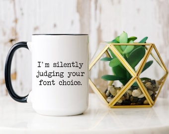 Puede incluir: Taza de cerámica blanca con asa y borde negros. La taza presenta el texto "I'm silently judging your font choice". Un terrario geométrico con una planta suculenta artificial se encuentra junto a la taza.