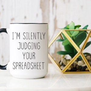 Könnte beinhalten: Weißer Keramikbecher mit schwarzem Henkel und Rand. Der Becher trägt den Text "I'M SILENTLY JUDGING YOUR SPREADSHEET" in Schwarz, zentriert auf der Vorderseite. Rechts befindet sich ein dekoratives geometrisches Terrarium mit einer künstlichen Sukkulente.