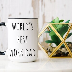 Può includere: Tazza in ceramica bianca con manico e bordo neri, con la scritta "WORLD'S BEST WORK DAD". Accanto alla tazza c'è un terrario geometrico in vetro con una pianta succulenta finta.