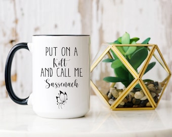 May include: White ceramic mug with black handle and rim, featuring the text "PUT ON A KILT AND CALL ME Sassenach" in black script, along with a butterfly graphic. A decorative geometric terrarium with a green plant is next to the mug.