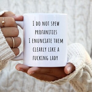 Könnte beinhalten: Ein weißer Keramikbecher mit schwarzem Aufdruck, der folgenden Text enthält: I DO NOT SPEW PROFANITIES I ENUNCIATE THEM CLEARLY LIKE A FUCKING LADY. Der Becher verfügt über einen Standardhenkel und eine glatte, glänzende Oberfläche für Getränke.