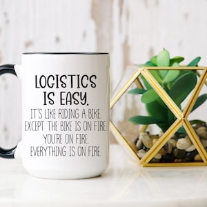 Può includere: Tazza in ceramica bianca con manico e bordo neri. La tazza presenta il testo "LOGISTICS IS EASY. IT'S LIKE RIDING A BIKE. EXCEPT THE BIKE IS ON FIRE. YOU'RE ON FIRE. EVERYTHING IS ON FIRE." Un terrario geometrico decorativo con una pianta è accanto alla tazza.