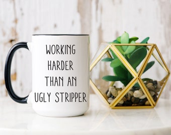 May include: White ceramic mug with a black handle and rim. The mug features the text "WORKING HARDER THAN AN UGLY STRIPPER" in black lettering. A geometric terrarium with a faux succulent is next to the mug.