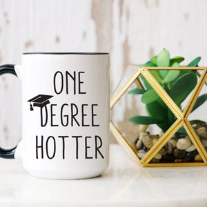 May include: White ceramic mug with black handle and rim, featuring the text "ONE DEGREE HOTTER" with a graduation cap graphic. A geometric glass terrarium with a green plant sits beside the mug.